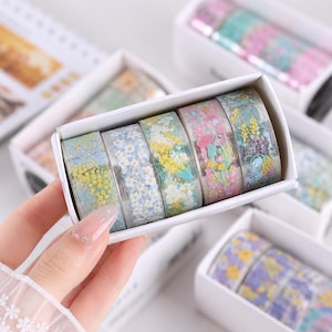 Könnte beinhalten: Ein Set aus fünf Rollen dekorativem Klebeband, angeordnet in einer weißen rechteckigen Schachtel. Jede Rolle zeigt ein eigenes Blumenmuster mit Farben wie Gelb, Blau, Weiß, Rosa und Grün. Die Bänder haben eine halbtransparente Oberfläche.