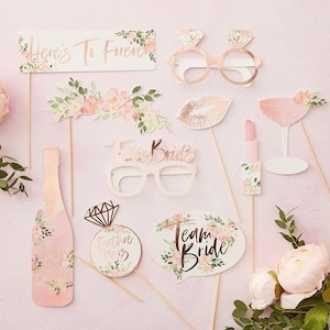 May include: A collection of pink and gold photo booth props for a bridal shower. Includes glasses, lips, a champagne bottle, and signs that say "Here's to Forever", "Future Mrs", and "Team Bride". Floral accents decorate the props.