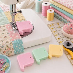 May include: A white sewing machine with a pink seam guide attached, alongside additional pink, mint green, and yellow seam guides. The setup includes spools of thread, a rotary cutter, a pin cushion, and fabric clips for quilting projects.