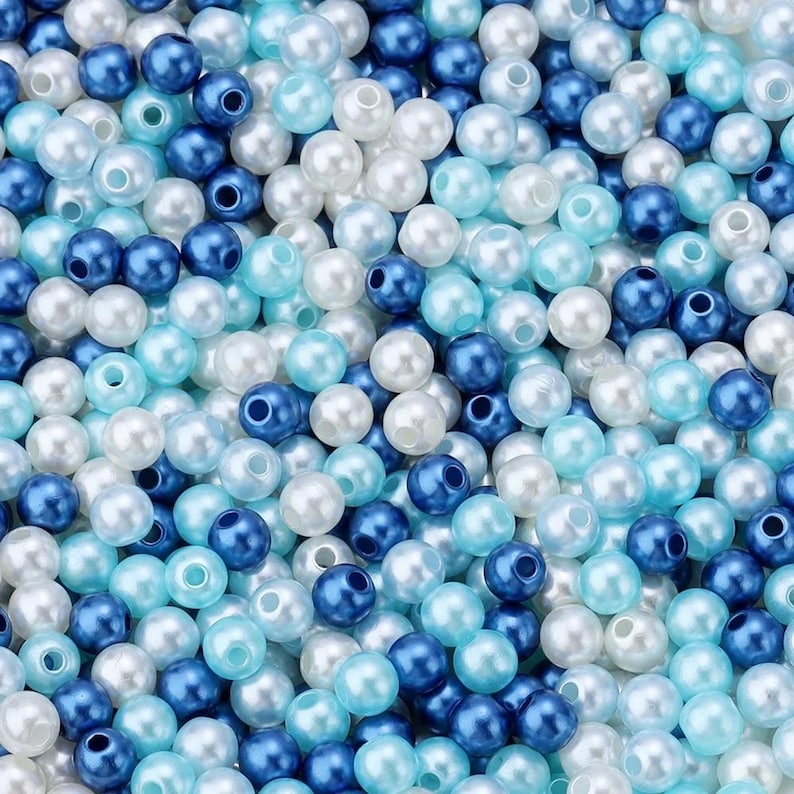 May include: A close-up shot of a pile of round beads in various shades of blue and white. The beads are scattered, creating a textured surface. The colors range from deep navy to light sky blue and pearly white, suggesting a nautical or ocean theme.