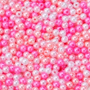 May include: A close-up shot of a pile of small, round beads in shades of pink, white, and peach. The beads have a glossy, pearl-like finish and are scattered randomly, creating a textured surface. These beads are ideal for jewelry making and crafts.