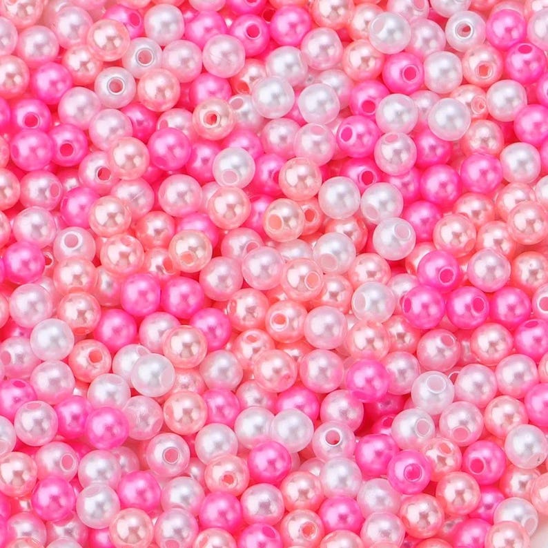 May include: A close-up shot of a pile of small, round beads in shades of pink, white, and peach. The beads have a glossy, pearl-like finish and are scattered randomly, creating a textured surface. These beads are ideal for jewelry making and crafts.