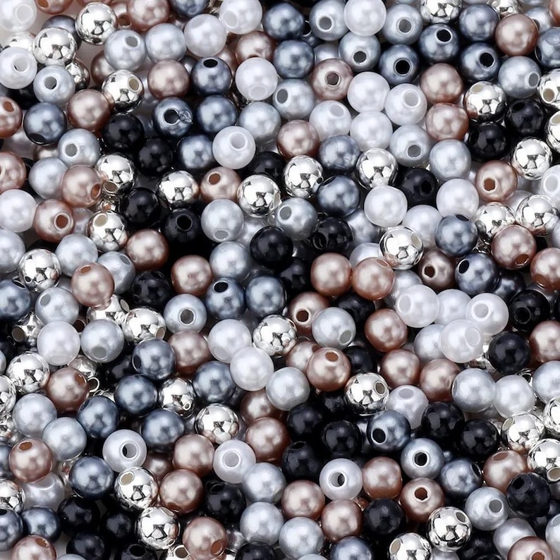 May include: A close-up shot of a collection of beads in various colors. The beads are a mix of pearlized white, silver, black, gray, and bronze. The beads are round with a hole through the center, and are scattered randomly.