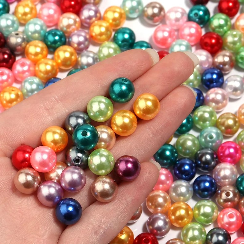 May include: A collection of colorful, round beads in various shades of red, pink, blue, green, gold, and pearl. The beads are scattered, with some held in a hand, showcasing their glossy, smooth surfaces. These beads are suitable for jewelry making and crafts.