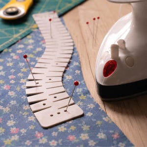 May include: A white plastic sewing seam guide with multiple slots and holes, pinned onto blue fabric with a floral pattern. A white iron with a red indicator light sits nearby, alongside a yellow rotary cutter on a green cutting mat.