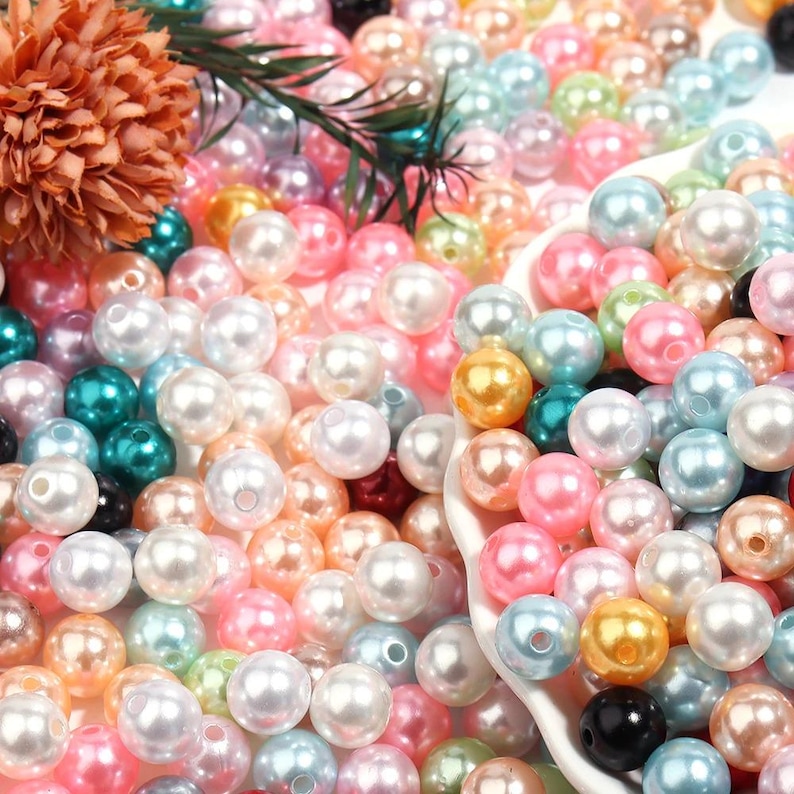 May include: A close-up shot of a collection of colorful beads in various shades of pink, white, gold, teal, and blue. The beads are round and have a glossy finish. A few decorative flowers and leaves are visible in the background.