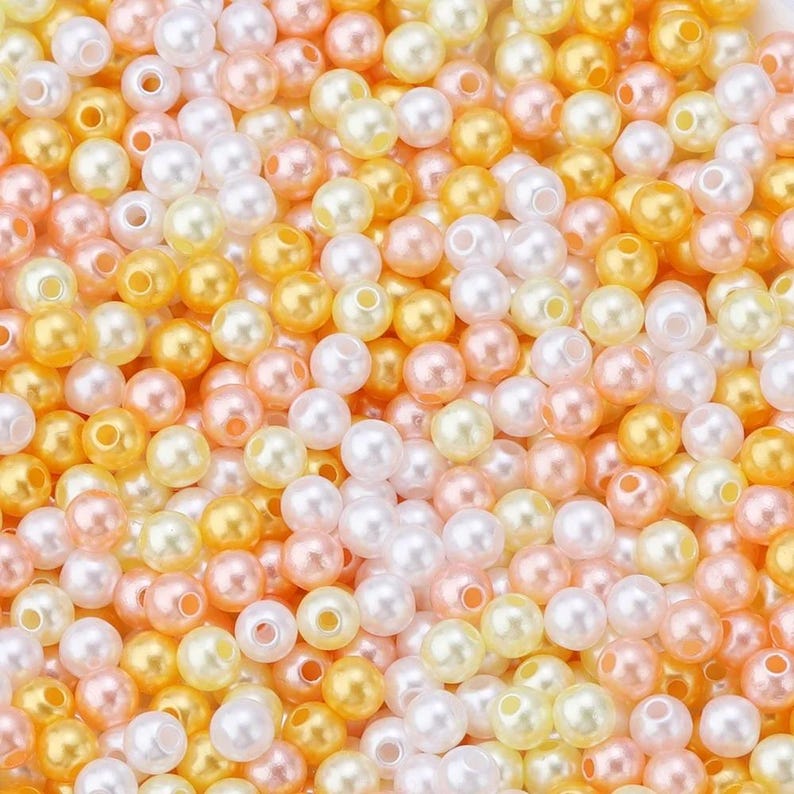 May include: A close-up view of a pile of small, round beads in various colors. The beads are a mix of white, peach, yellow, and gold, creating a warm and inviting palette. These beads are likely used for jewelry making or other craft projects.