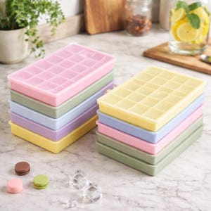 Può includere: Pile di vassoi per cubetti di ghiaccio in silicone colorati in tonalità pastello rosa, verde, blu e giallo. I vassoi sono rettangolari con più scomparti quadrati. L'immagine include anche macarons e cubetti di ghiaccio trasparenti.