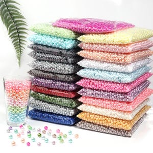 May include: A collection of colorful pearl beads in various shades, including pink, green, blue, and white, are displayed in clear plastic bags. A glass filled with gradient-colored beads sits to the left, with loose beads scattered in front. These beads are ideal for crafting.