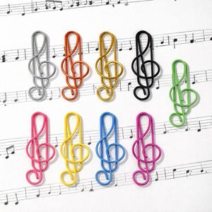 May include: A selection of vibrant metal paper clips in the shape of treble clefs. The clips are in a range of colours: silver, orange, gold, black, green, pink, yellow, and blue, displayed on a sheet of musical notation.