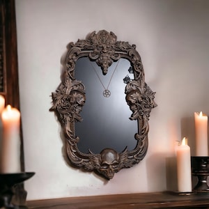 Può includere: Specchio con cornice marrone ornata con abbellimenti a forma di teschio e floreali. Lo specchio ha una superficie scura e riflettente e un ciondolo con un simbolo a stella. La cornice presenta intagli dettagliati e un teschio alato in basso.