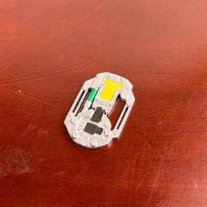 May include: A small, silver-coloured metallic token with a rectangular shape and rounded edges. The surface features a central yellow rectangular inlay, a thin green accent line, and a black printed circuit board design with intricate line details.