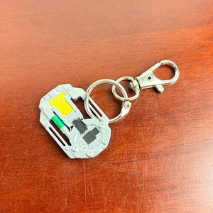 May include: A silver-toned metal keyring featuring a circular, mechanical-style pendant with a yellow rectangular accent, a small green cylindrical detail, and black circuitry-like markings, attached to a split ring and a lobster claw clasp.