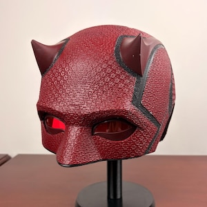 Op de afbeelding: Een getextureerde donkerrode helm met twee prominente hoorns aan de bovenkant en donkerrode ooglenzen. De helm is gemonteerd op een zwarte ronde standaard. Het oppervlak van de helm heeft een duidelijk herhalend patroon en zwarte lijnen.