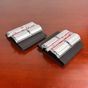 May include: Two silver metal objects with red detailing are attached to black elastic bands. The rectangular objects have multiple sections. The items are displayed on a dark wooden surface.