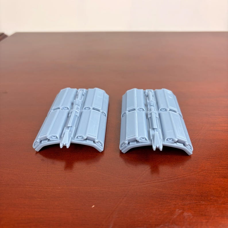 May include: Two light gray, curved, rectangular objects with a series of rectangular panels and a central structural element. The objects are placed side by side on a brown surface. The objects appear to be components or parts of a larger item.