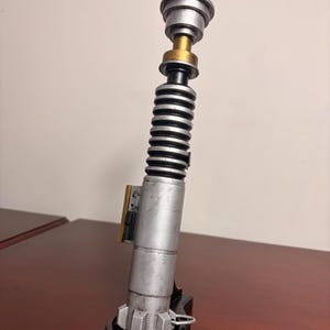 May include: A silver and black lightsaber prop with gold accents. The prop has a cylindrical body with a ribbed section and a detailed top. The lightsaber is a replica of a popular science fiction weapon.