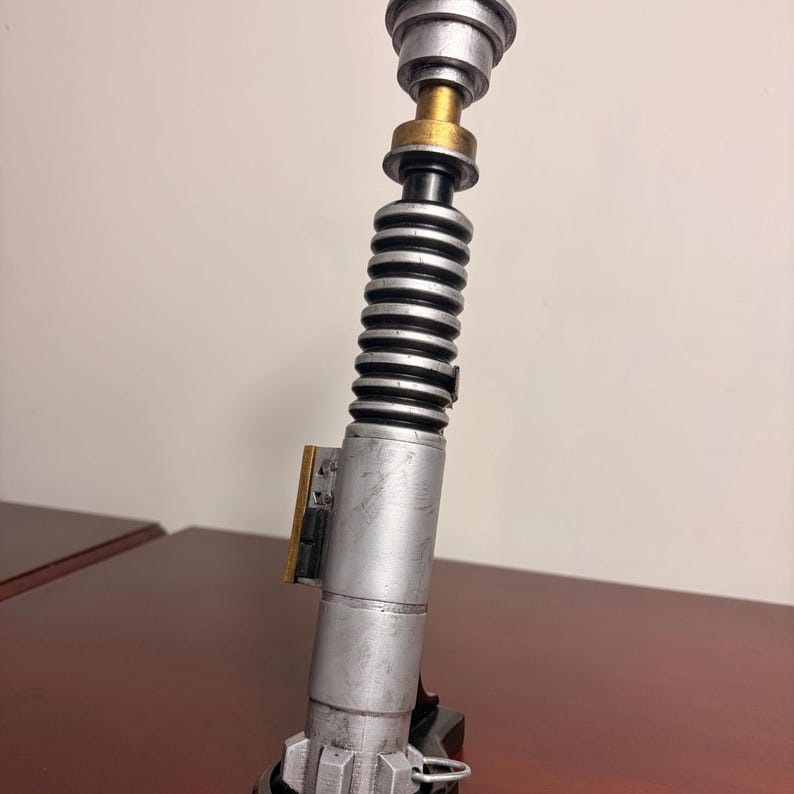 May include: A silver and black lightsaber prop with gold accents. The prop has a cylindrical body with a ribbed section and a detailed top. The lightsaber is a replica of a popular science fiction weapon.