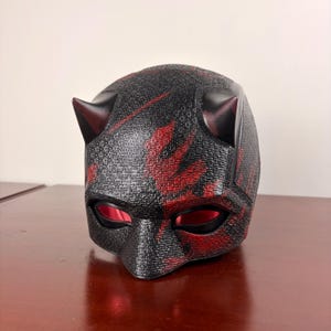 May include: A black and red textured mask with two small horns on top. The mask has red accents and eye openings. The mask is likely a costume or cosplay item.