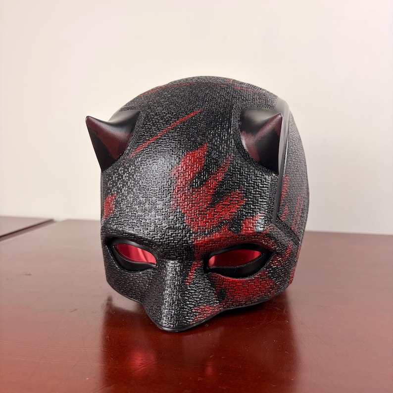May include: A black and red textured mask with two small horns on top. The mask has red accents and eye openings. The mask is likely a costume or cosplay item.