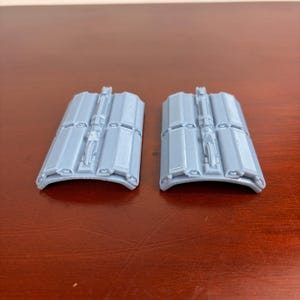 May include: Two light gray, curved, rectangular objects with detailed ridges and a central structure. The objects are symmetrical and appear to be components of a larger structure or device. They are placed on a dark brown surface.