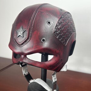 May include: A burgundy helmet with a distressed finish, a silver star emblem, and a textured side panel. The helmet has eye openings and a chin strap with white straps.