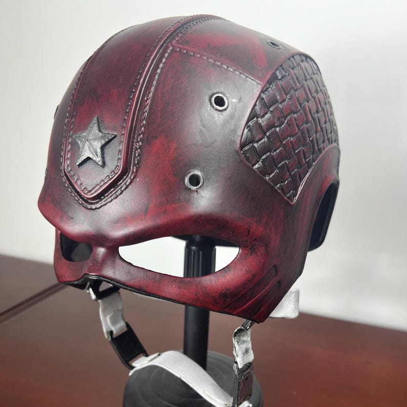 May include: A burgundy helmet with a distressed finish, a silver star emblem, and a textured side panel. The helmet has eye openings and a chin strap with white straps.