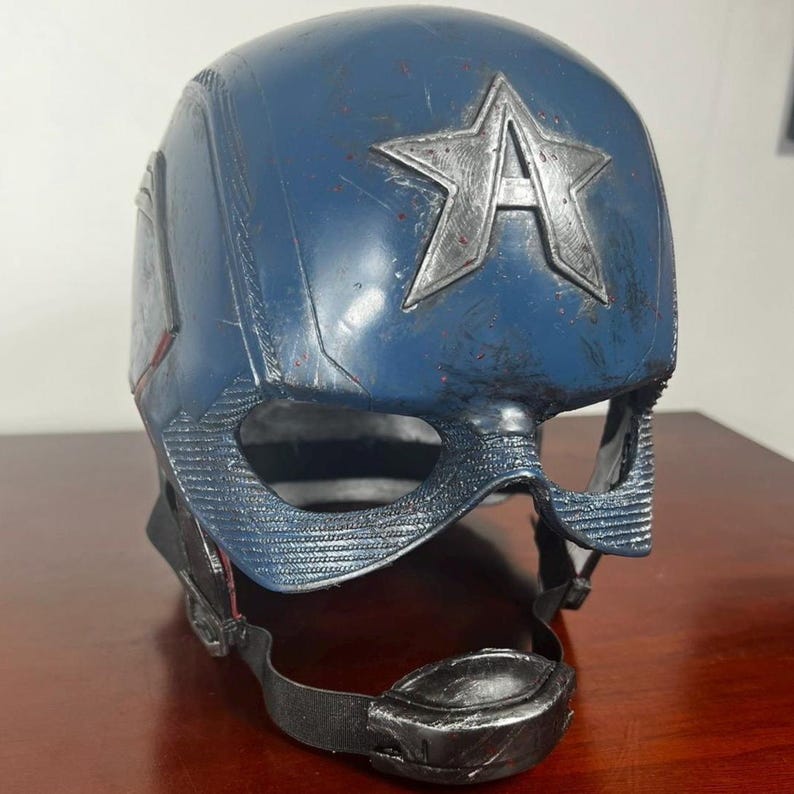 May include: A blue and silver Captain America helmet. The helmet has a large silver star with the letter "A" in the centre. It features a chin strap and eye protection. The helmet is made of a durable material and is designed to protect the wearer's head.