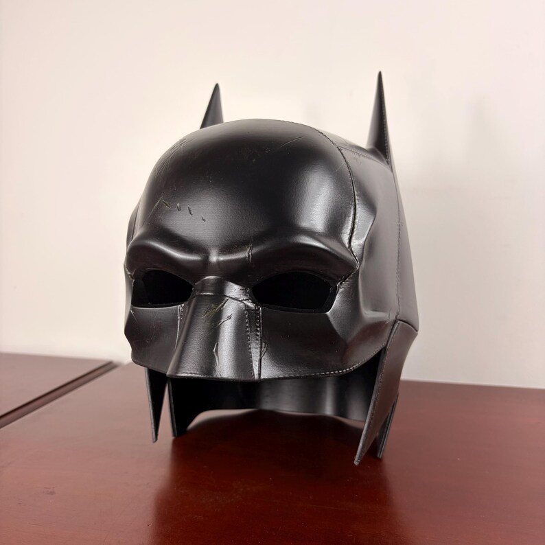 May include: A black Batman cowl with pointed ears and a textured finish. The mask features eye openings and a defined nose and chin, with visible stitching and subtle wear marks. The design is sleek and detailed.