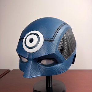 Op de afbeelding: Een blauwe helm met een opvallend wit cirkelvormig doelwit-embleem op het voorhoofd. De helm heeft een getextureerd oppervlak, zwarte zijpanelen met een rasterpatroon en twee oogopeningen. Het is gemonteerd op een zwarte standaard.