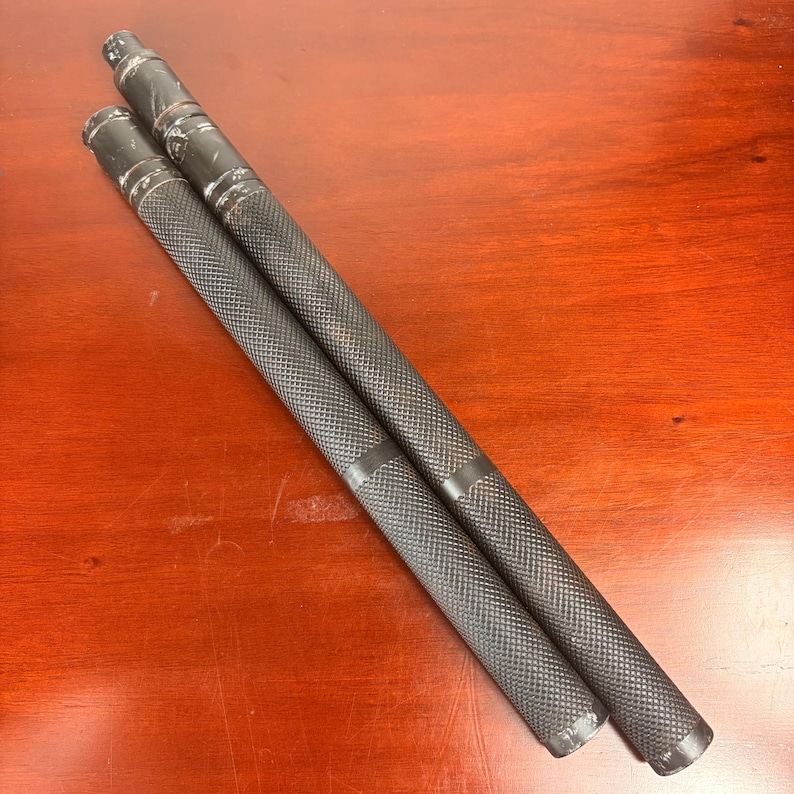 May include: Two black metal bars with textured grips are displayed diagonally on a wooden surface. The bars have a cylindrical shape with a diamond pattern on the grip area. The ends of the bars have a different design, with a series of rings.