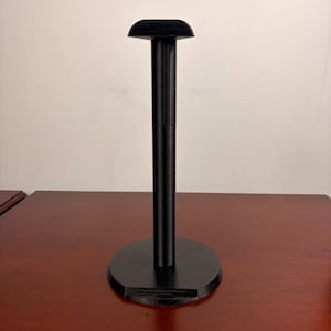 May include: A black headphone stand featuring a circular base, a vertical cylindrical support column, and a curved top rest piece designed to hold a pair of over-ear headphones. The stand has a matte finish and a simple, functional structure.