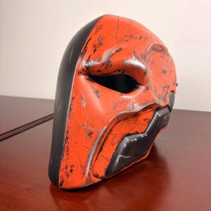 May include: An orange and black mask with a distressed finish, showing signs of wear and tear. The mask has a distinctive eye opening and angular features. The surface is marked with scratches, giving it a used appearance. Likely a costume or cosplay item.