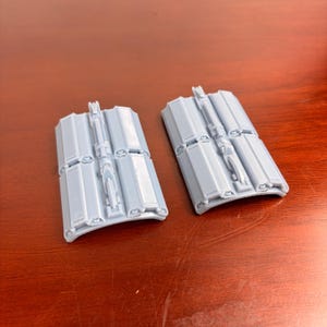 May include: Two light gray, rectangular plastic components with a series of rectangular panels and a central, raised, vertical structure. The components have a curved bottom edge and are set against a brown surface.