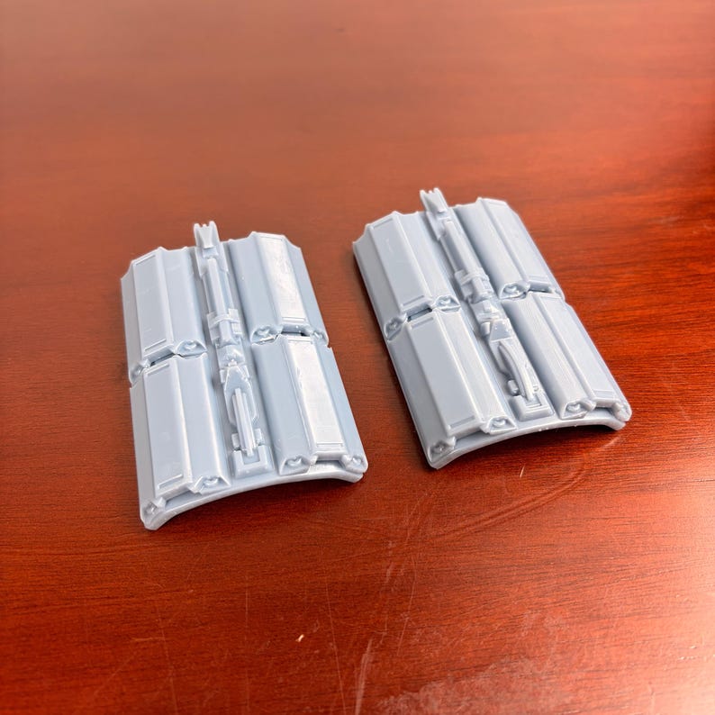 May include: Two light gray, rectangular plastic components with a series of rectangular panels and a central, raised, vertical structure. The components have a curved bottom edge and are set against a brown surface.