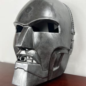 May include: A silver-coloured mask with a distinctive angular design. The mask features a prominent nose, a rectangular mouth opening, and eye openings. The surface has a weathered appearance, suggesting a vintage or battle-worn aesthetic. The mask is likely a costume or collectible item.