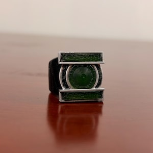 Puede incluir: Un anillo que presenta un emblema circular verde central con un patrón de círculos concéntricos, flanqueado por dos barras rectangulares verdes horizontales. El diseño está encerrado en un marco de metal plateado con una banda negra.