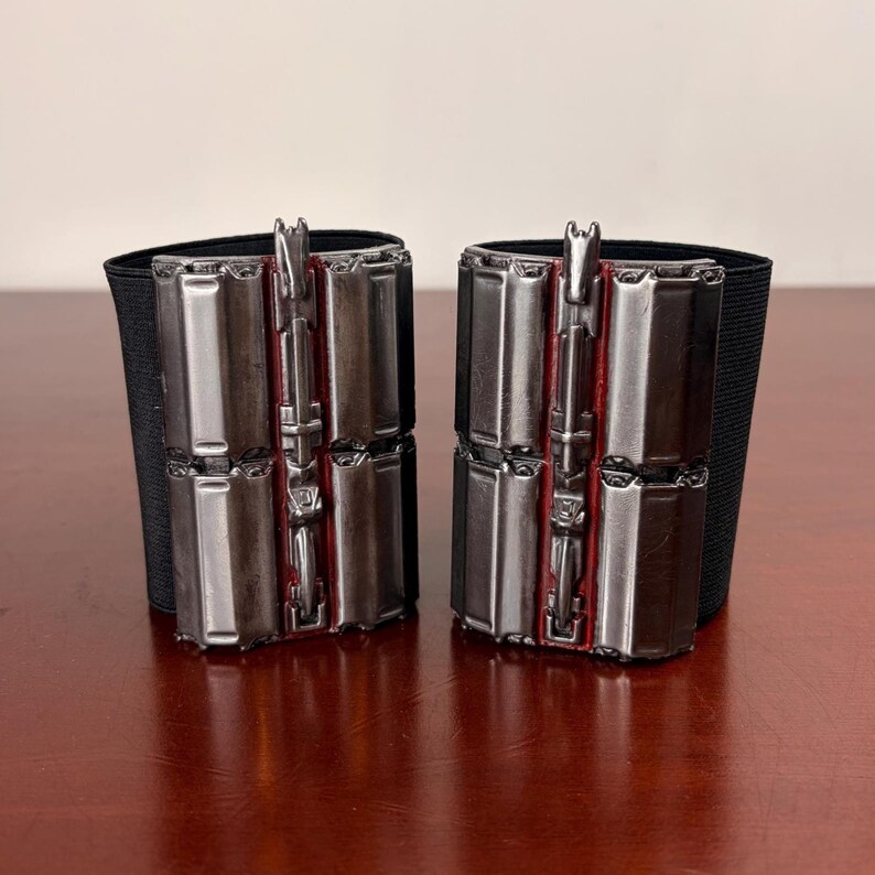 May include: A pair of silver-coloured wrist cuffs, each featuring a series of rectangular metal plates and red detailing. The cuffs are attached to a black elastic band. The cuffs are displayed on a wooden surface.
