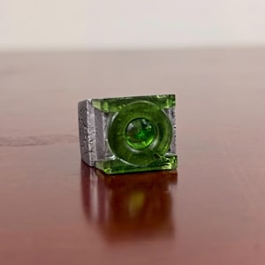 May include: A silver-toned ring featuring a square-shaped face with a textured, weathered metal band. The centre of the ring displays a translucent, circular green gemstone element set within a raised, square-shaped green frame.