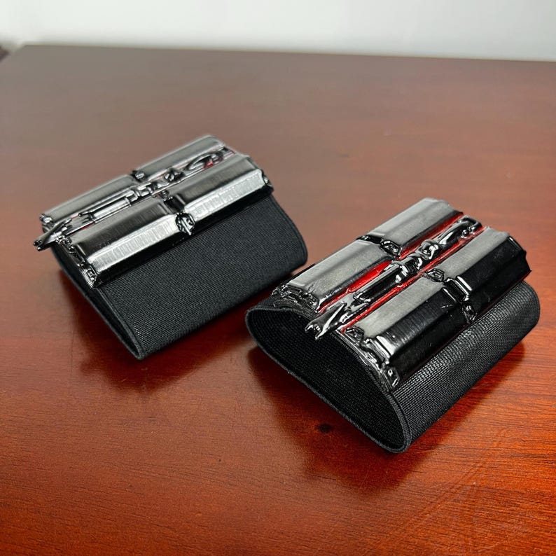 May include: A pair of black wrist cuffs with a rectangular design. The cuffs have a shiny, metallic appearance with a red accent. The cuffs are attached to a black elastic band.
