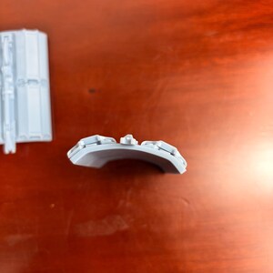 May include: Two light gray plastic components. One is a rectangular piece with a series of parallel lines and a few protruding elements. The other is a curved piece with several raised, rectangular details.