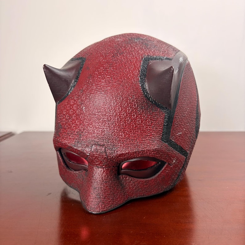 May include: A red textured mask with two dark brown horns on top. The mask has eye openings with red lenses and black accents. This mask is likely a costume or cosplay item.