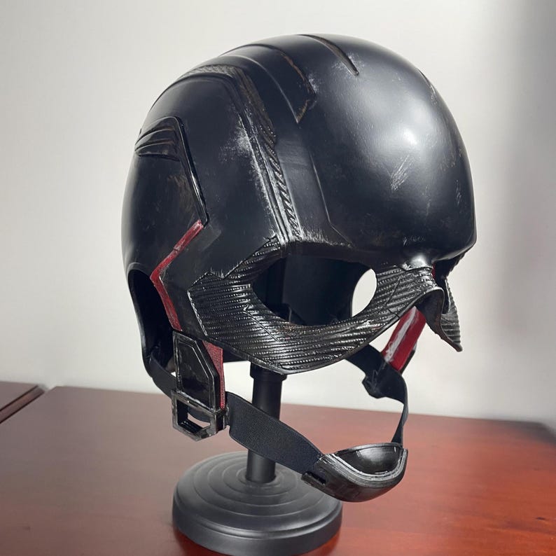 May include: A black helmet with red detailing, presented on a black stand. The helmet features a textured surface with visible lines and details. The design includes a chin strap and a visor area. The helmet appears to be a prop or costume piece.