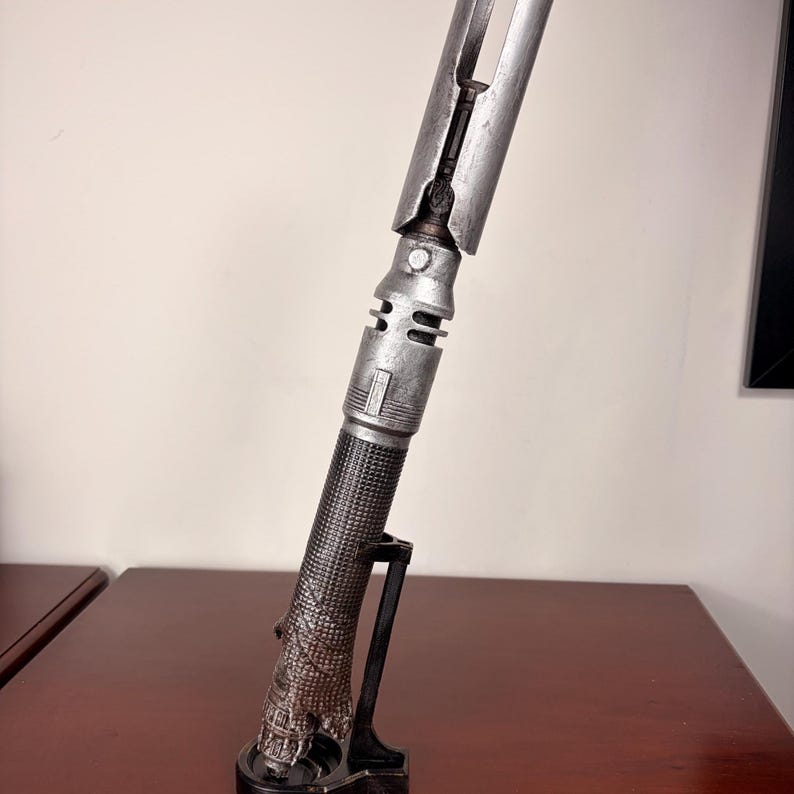 May include: A silver and black lightsaber prop, displayed on a black stand. The lightsaber has a detailed, textured handle and a metallic blade. The prop is designed to resemble a futuristic weapon.