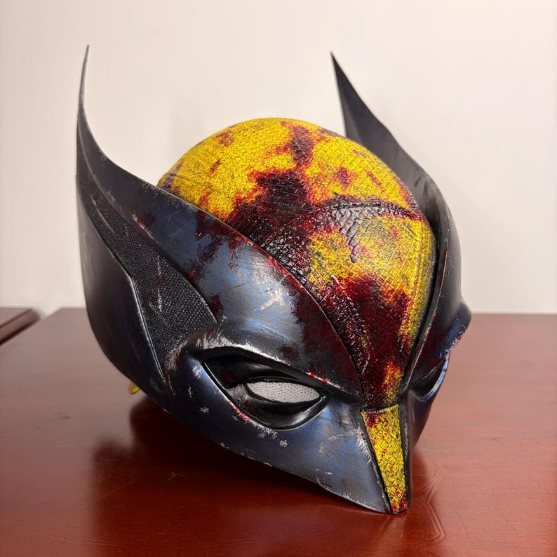 May include: A Wolverine-style mask with a yellow and red patterned top and dark blue accents. The mask has pointed ears and eye openings. The mask is made of a hard material, likely a costume accessory.