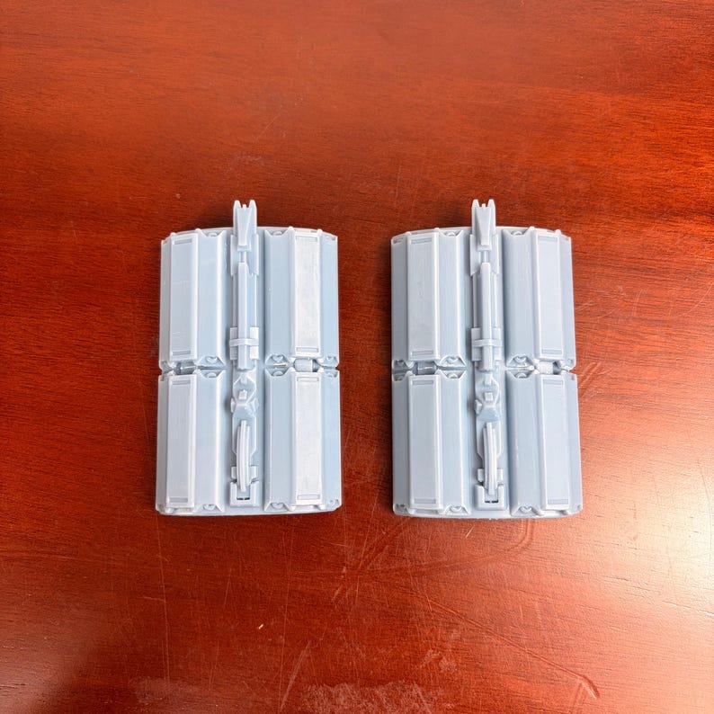 May include: Two light gray, plastic, mechanical components. Each component has a rectangular shape with multiple hinged sections and a central, vertical structure with intricate details. The components are designed to fit together.