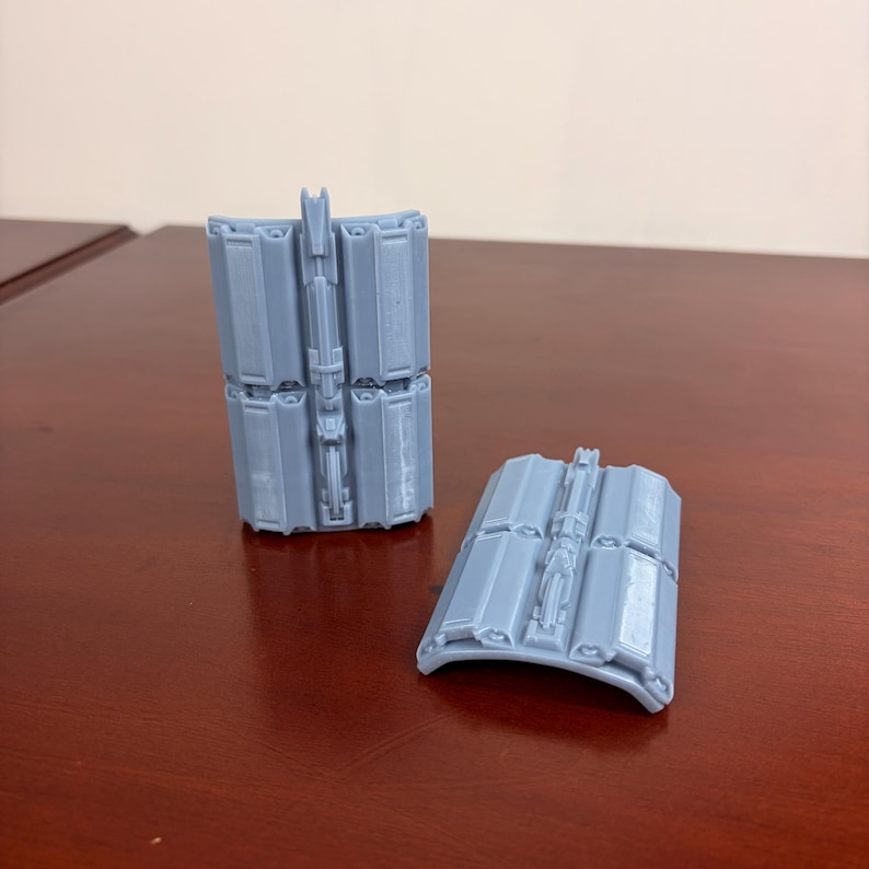 May include: Two light gray, 3D-printed objects. One is upright, the other is lying down. Both have a curved shape and a series of rectangular panels. The objects appear to be components of a larger structure or device.