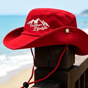 May include: A bright red wide-brimmed hat featuring a white embroidered mountain graphic and the text "Outdoor Lifestyle" on the front. The hat includes a matching red chin strap with an adjustable black toggle and a small metallic side button.