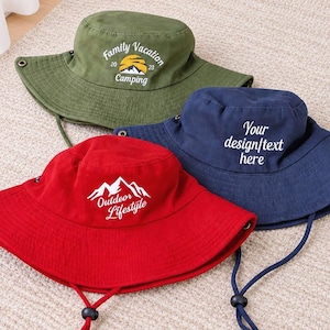 May include: Three bucket hats in olive green, navy blue, and red. The olive hat features a mountain graphic with text reading Family Vacation 2023 Camping. The navy hat has text reading Your design/text here. The red hat shows a mountain graphic with text reading Outdoor Lifestyle.