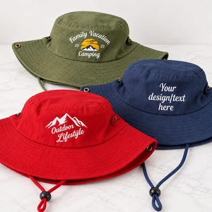 May include: Three wide-brimmed bucket hats in olive green, navy blue, and red. The olive hat features a sunset and mountain graphic with text reading Family Vacation 2023 Camping. The red hat displays a mountain graphic and Outdoor Lifestyle text.